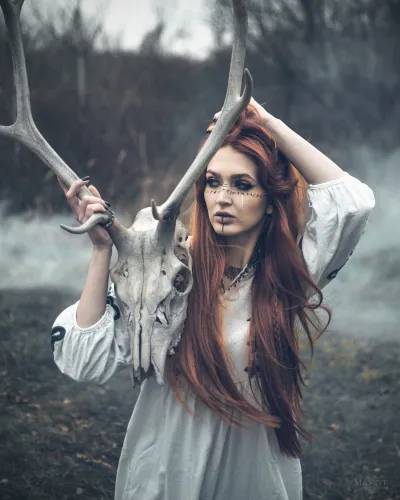 Artistic portrait of a woman with red hair holding a deer skull in a mystical setting